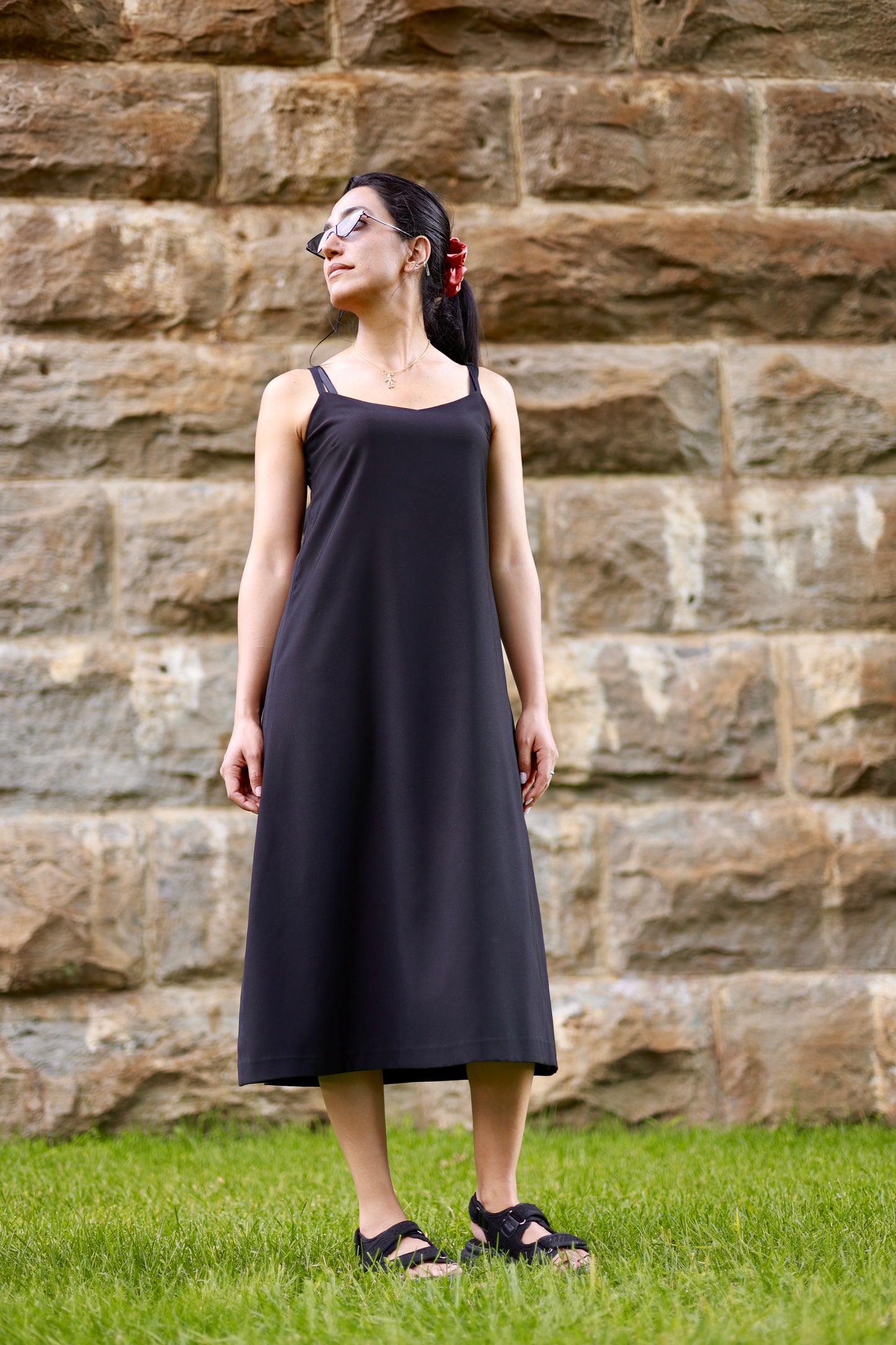 Robe noir en soie par Couturière Ile des Soeurs ELZi. Photo prise à Sainte-Anne-de-Bellevue. Montreal Dressmaker ELZi wears the black silk dress with black sunglasses in front of a stone background.