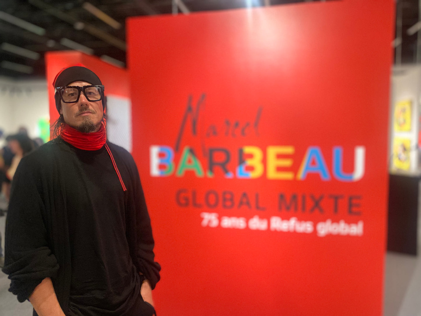 Jonathan Brunelle devant l'accueil de l'exposition Marcel Barbeau : Global mixte au Musée des beaux-arts de Mont-Saint-Hilaire pour célébrer les 75 ans de la parution du Refus global. Il porte un cache cou rouge en laine merino par ELZi.ca
